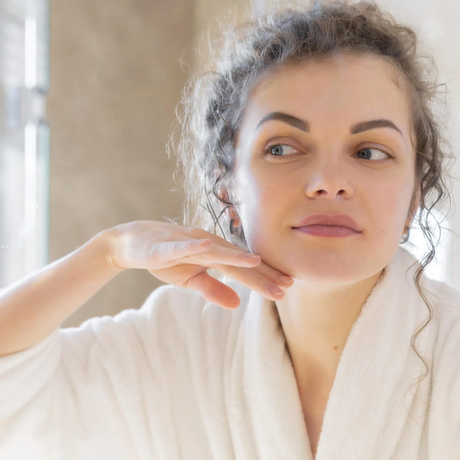 Woman examining her skin texture on her cheek near a window